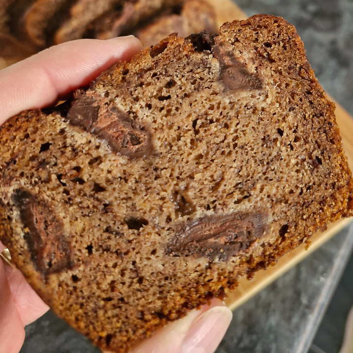 A close-up of a hand holding a thick slice of chocolate chip banana bread, showing the moist brown crumb and dark chocolate chunks, with more sliced bread in the background.