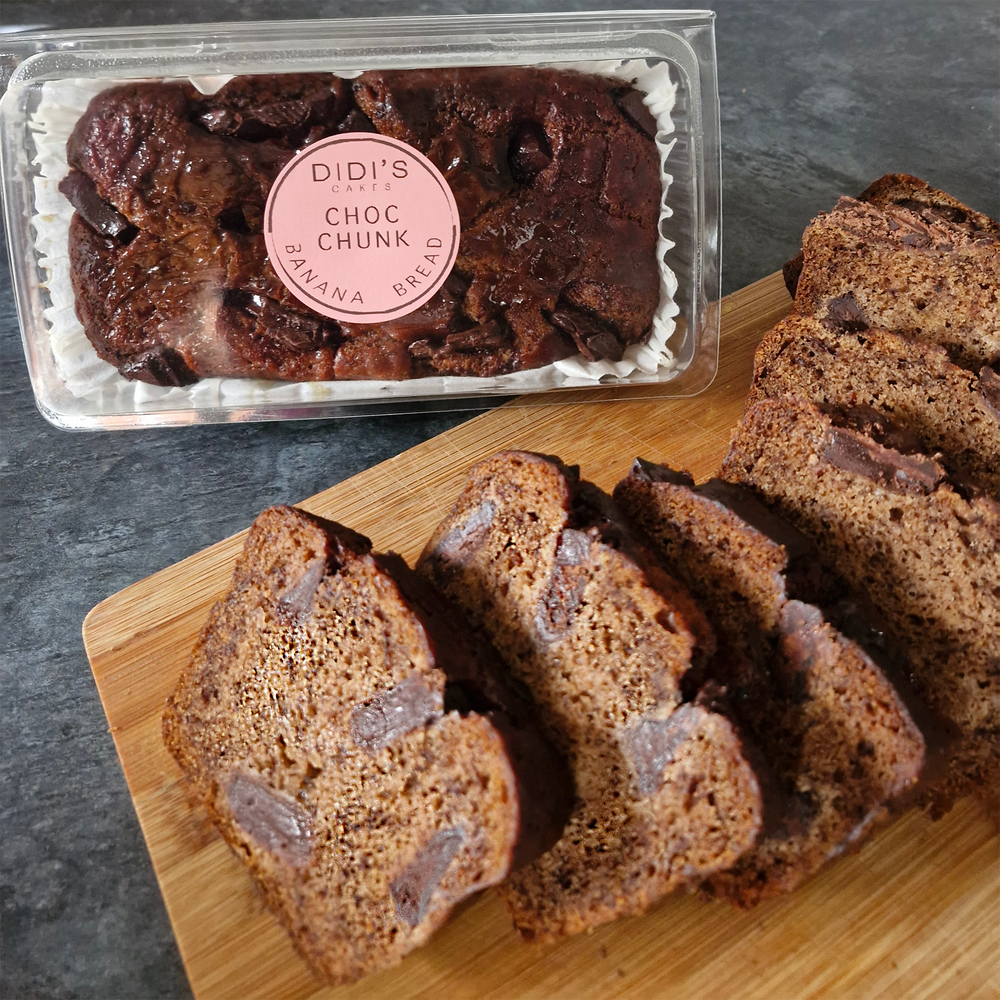 Chocolate chunk banana bread shown as a sliced loaf on a wooden cutting board next to its clear plastic packaging container with a pink circular label reading 'DIDI'S CHOC CHIP BANANA BREAD'.