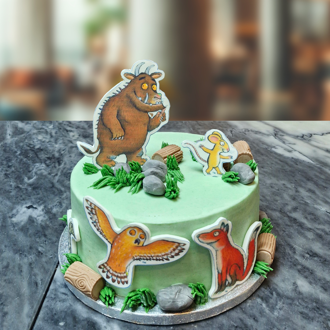 A decorated cake with characters from the Gruffalo storybook, including a Gruffalo, fox, and owl, presented on a plate against a blurred background.