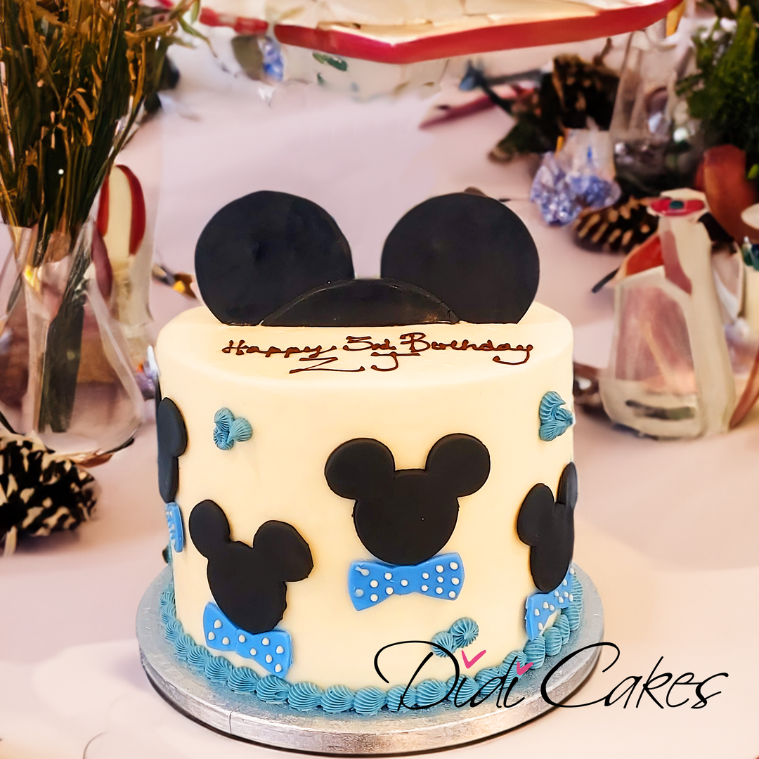A themed birthday cake with Mickey Mouse design, featuring layers of white icing and black decorations, with a happy birthday message, and a small Mickey Mouse figure on top.