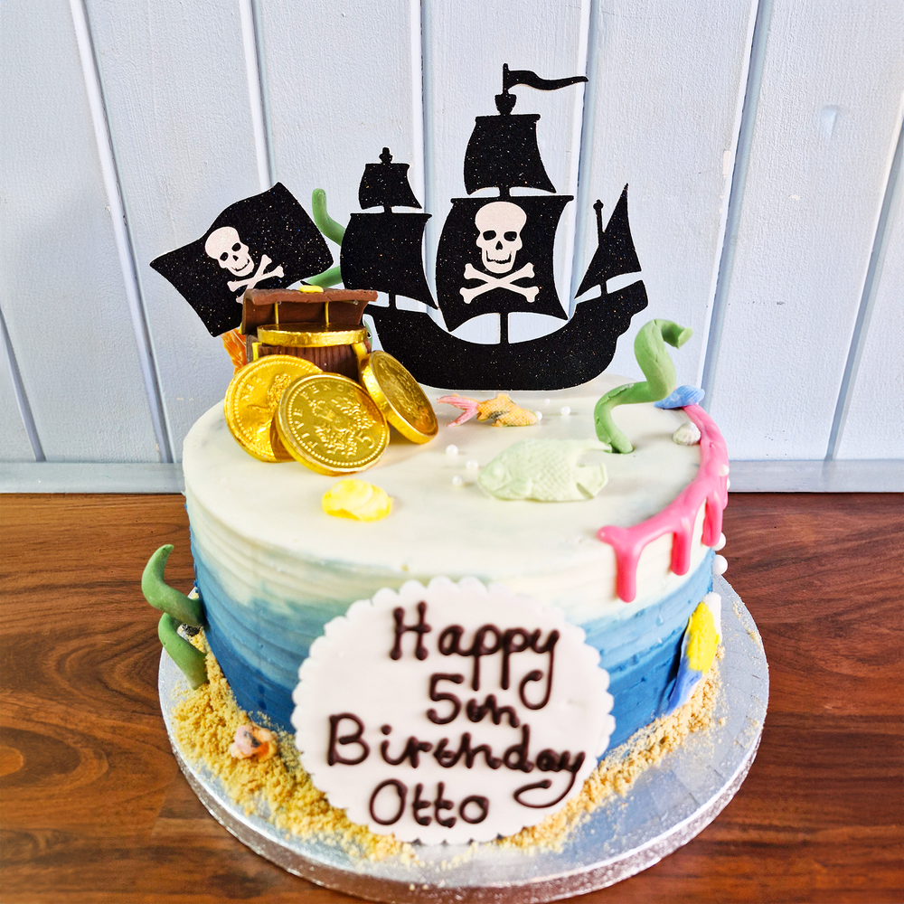 Pirate-themed birthday cake with ship topper and 'Happy 5th Birthday Otto' message on a wooden surface.