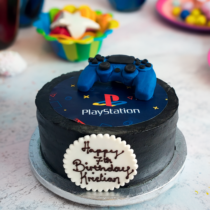 A round birthday cake decorated with a PlayStation logo and a blue playstation controller on top, with a &